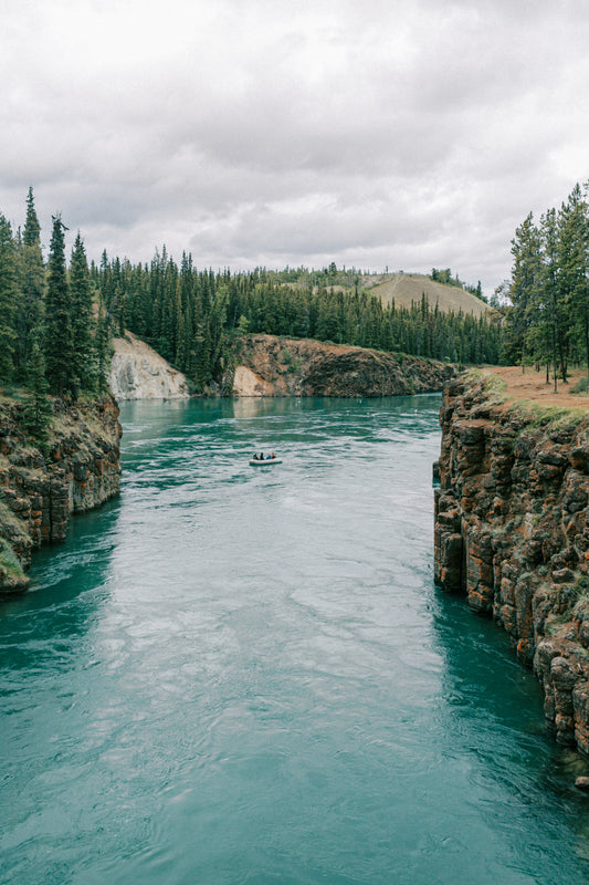 A small boat floats on a clear blue river surrounded by a lush forest. The river flows between rocky cliffs, with tall evergreen trees lining the banks. The sky above is cloudy, casting a serene and tranquil atmosphere on the landscape.