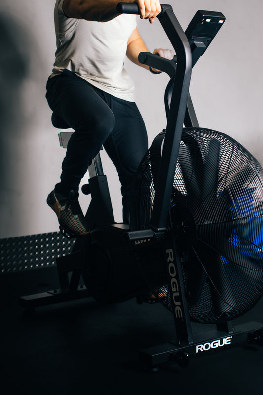 A person wearing a white shirt and black pants works out on a stationary exercise fan bike in a dimly lit room. The bike is branded "Rogue" and has a large fan wheel at the front. Only the person's torso and legs are visible.