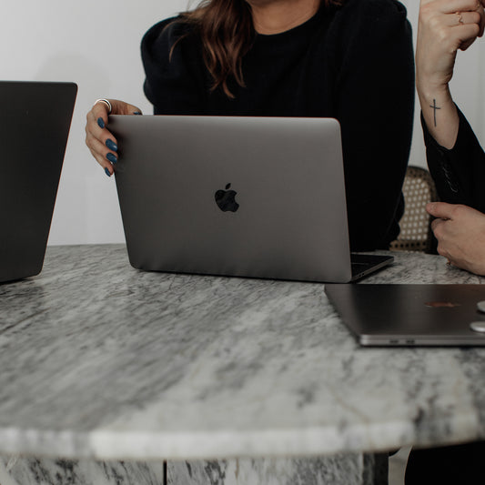 Two people sit at a marble table with laptops. One person, with dark hair and blue-painted nails, holds a gray Apple laptop. The other person, whose hand has a cross tattoo, is partially visible next to another laptop on the table.