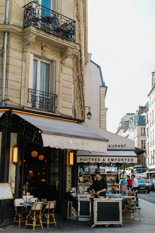 A street scene of a French café with tables and chairs set up outside under a striped awning. The surrounding buildings display classic Parisian architecture with wrought-iron balconies. A man is working at a seafood stall labeled "Huîtres à Emporter".