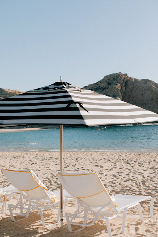 A beach scene with two white lounge chairs under a large black and white striped umbrella. The sandy shoreline leads to calm, clear blue water, with rugged cliffs visible in the background under a clear, blue sky.