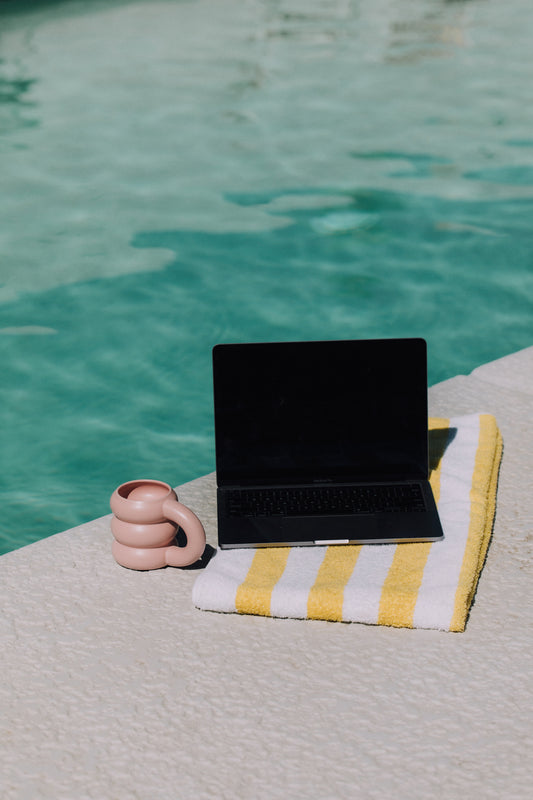 A black laptop rests on a yellow and white striped towel beside a clear swimming pool. Next to the laptop is a unique pink mug with an artistic, curvy handle. The scene captures a blend of relaxation and productivity by the poolside.