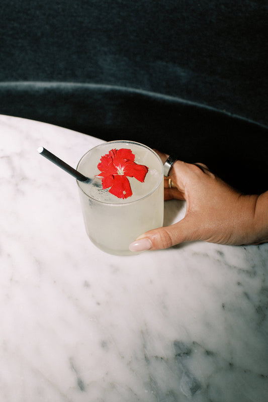 A hand wearing a silver ring holds a cocktail glass with a straw, sitting on a marble table. The drink is garnished with a bright red flower.