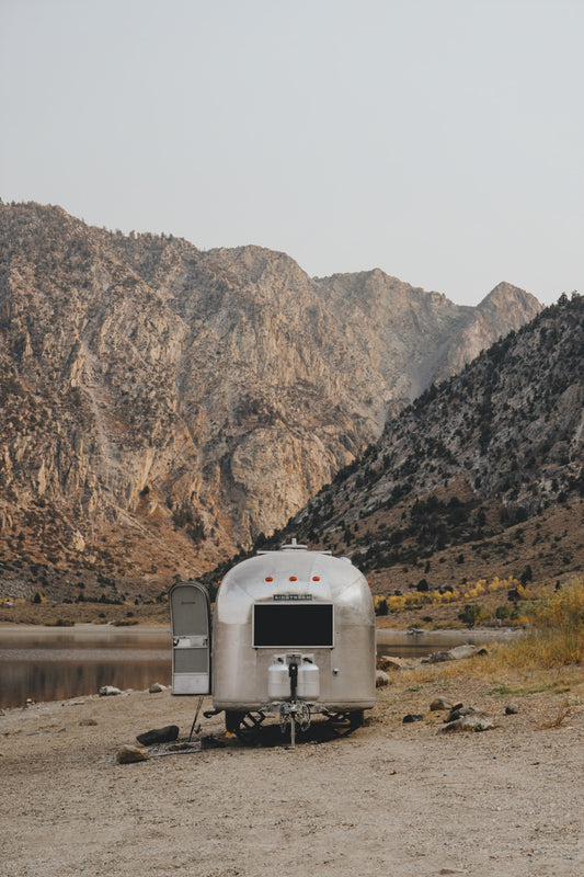 A silver camper trailer is parked by a calm lake surrounded by towering, rocky mountains. The trailer's door is open, and the landscape features sparse vegetation, highlighting the rugged and serene natural environment.