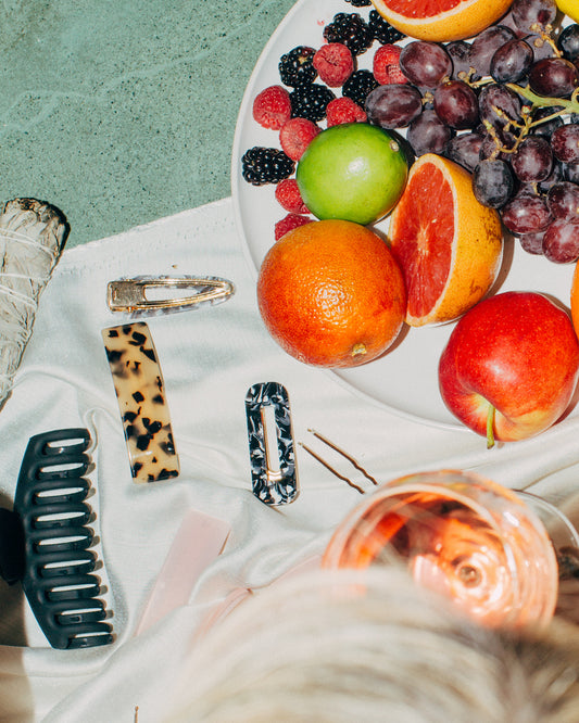 A plate of assorted fruits including grapes, oranges, limes, and raspberries sits on a white fabric. Nearby are various hair accessories, such as a black hair clip, a tortoiseshell hair clip, and a white hair clip, along with a glass of rosé.