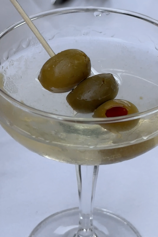 A close-up of a martini in a clear glass, garnished with three green olives on a cocktail pick. The drink has a pale, translucent appearance, and the glass sits on a white surface, partially visible. One of the olives has a red pimento stuffing.